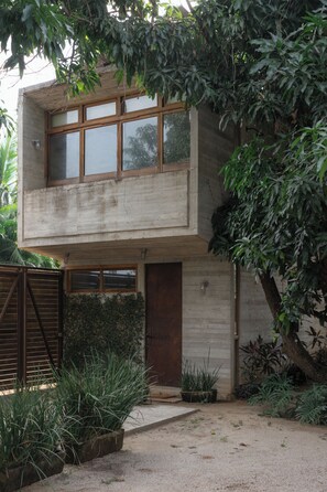 A two-story concrete building with large windows, a wooden door, and a small garden area with plants.