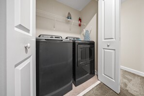 A laundry room with two front-loading washers and dryers, a fire extinguisher on the wall, and a shelf above the dryers. - 2921 SS - Storey Lake Dreamscape (Kissimmee)