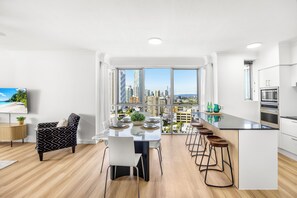 A modern kitchen with a large island, bar stools, and a dining area with a view of the cityscape. - Chevron Renaissance Ocean & River Views (Surfers Paradise)