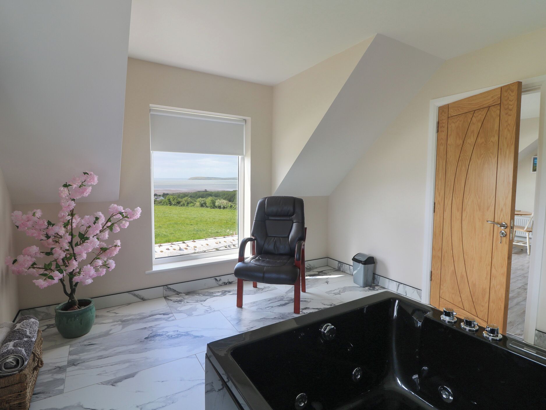 Una habitación con un ventanal grande, un sillón de cuero negro, una puerta de madera y una bañera de hidromasaje negra.