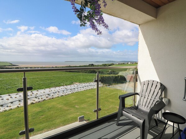 A balcony with a chair and table, overlooking a grassy area with a view of the sea and distant hills.
