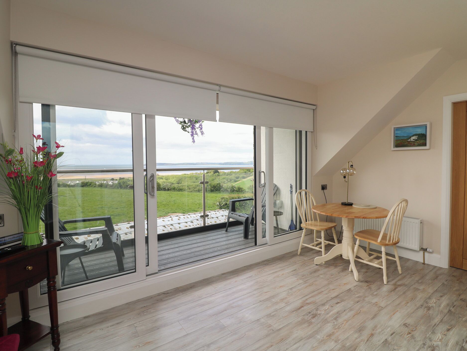 A room with a large sliding glass door leading to a balcony with a view of the sea. There is a wooden table with chairs and a small plant on the windowsill.