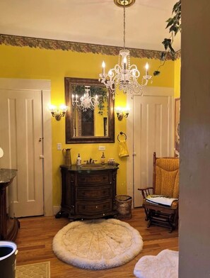 A room with a chandelier, a mirror, a wooden dresser, a chair, and a round rug.