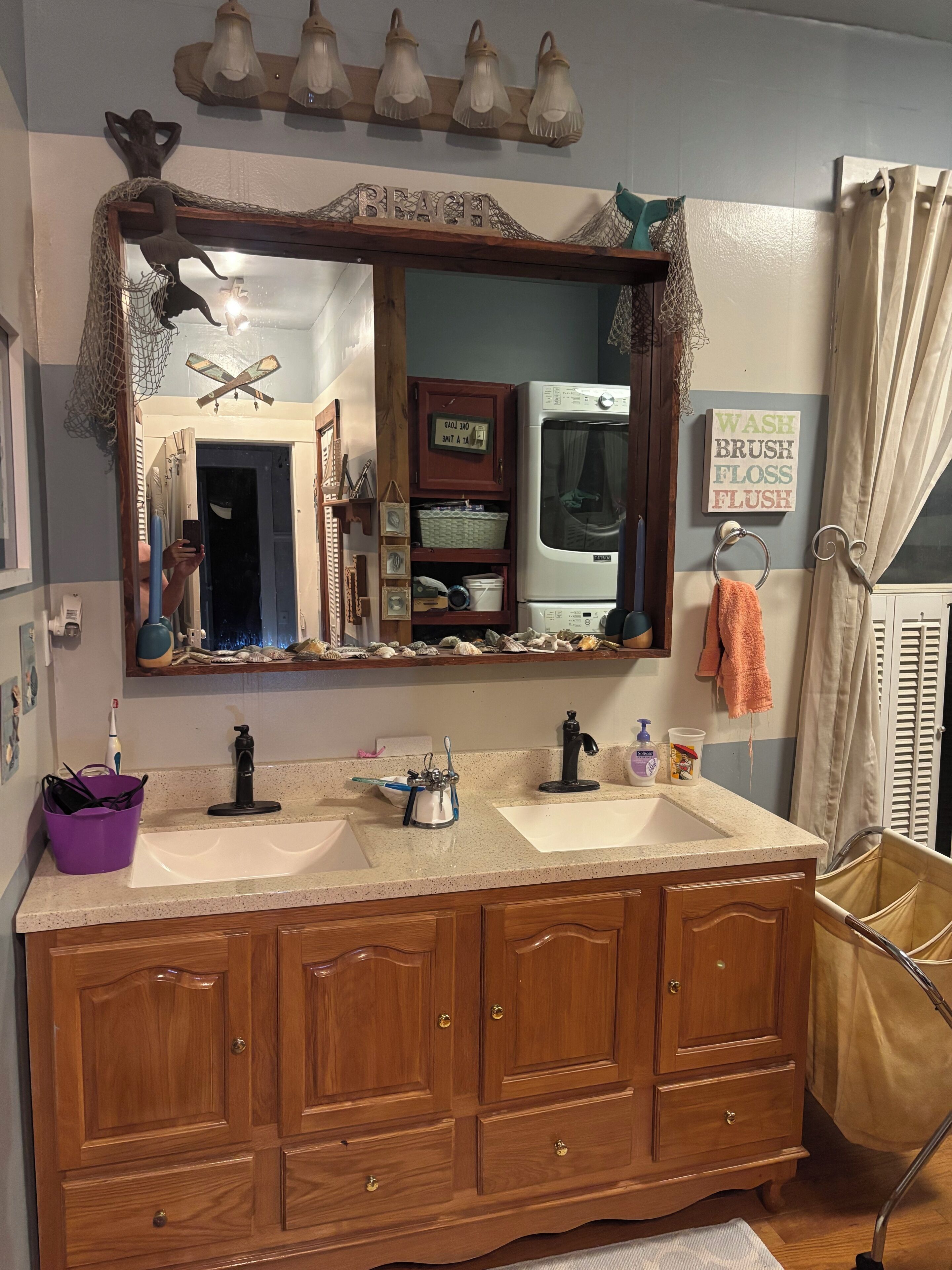 Un baño con tocador doble, espejo decorativo, perchero y carrito de lavandería.