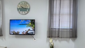 Une pièce avec une table en bois, deux chaises, un téléviseur diffusant une scène de plage, une horloge et une fenêtre ornée de rideaux légers.