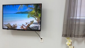 Un coin repas avec une table en bois, deux chaises grises, un téléviseur fixé au mur qui diffuse une scène de plage, une horloge et une orchidée en pot.