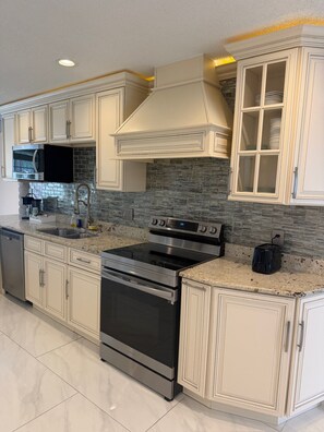 A kitchen with white cabinetry, a stainless steel oven, a microwave, and a stone backsplash. - The Marble Manor by Boardwalk (Myrtle Beach)