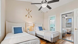 Et soveværelse med to senge, en ventilator i loftet, et natbord med lampe, og en dør til et badeværelse.