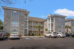 A multi-story building with a stone facade, balconies, and a parking lot with several cars. - Peaceful Point | Golf Course Views (Myrtle Beach)