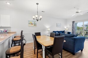 A modern living room with a blue sofa, wooden dining table, and chairs. - On Holiday | Pool Views + Private Balcony (Myrtle Beach)
