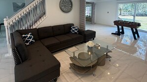 A spacious living room with a sectional sofa, a glass coffee table, a large wall clock, and a foosball table. - The Marble Manor | House & Loft (Myrtle Beach)