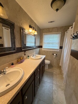 A bathroom with two sinks, a toilet, a window with blinds, and a shower curtain. - The Marble Manor | House & Loft (Myrtle Beach)