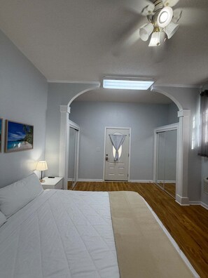 Um quarto com uma cama, uma mesinha de cabeceira, um abajur, um ventilador de teto e uma porta com janela.