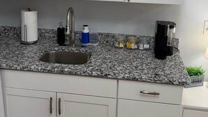 A kitchen with white cabinets, a granite countertop, a sink, a coffee maker, and a small potted plant. - The Marble Manor | House & Loft (Myrtle Beach)