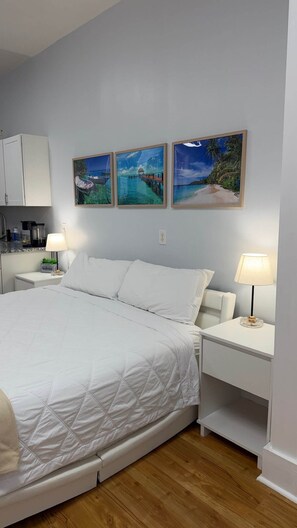 A hotel room with a bed, two bedside tables, a nightstand, a kitchen area, and three framed pictures on the wall. - The Marble Manor | House & Loft (Myrtle Beach)
