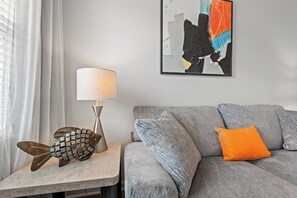 Ein Wohnzimmer mit grauem Sofa, einem orangefarbenen Kissen, einer Lampe und einer Fischskulptur auf einem Beistelltisch.