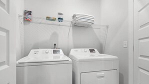 Une buanderie équipée de deux machines à laver et de deux sèche-linge à chargement frontal, d’une étagère en métal où sont pliées des serviettes, et de produits de lessive.