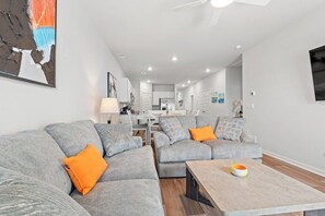 Ein modernes Wohnzimmer mit grauen Sofas, orangefarbenen Kissen, einem Couchtisch und einem Essbereich im Hintergrund.