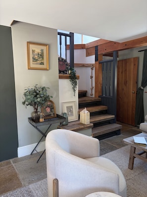 A living room with a sofa, a coffee table, a staircase with wooden railings, and a door with a curtain. - THE LOST RADISH - Cosy and romantic Cotswold cottage with private garden (Kingham)