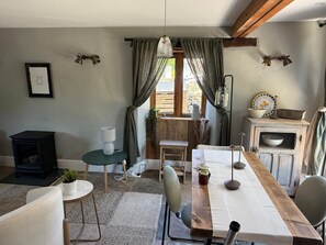 A cozy room with a wooden table, chairs, a small round table with a lamp, a fireplace, and a window with curtains. - THE LOST RADISH - Cosy and romantic Cotswold cottage with private garden (Kingham)