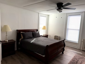 Une chambre à coucher comprenant un lit, deux tables de chevet, un ventilateur de plafond et une fenêtre avec des stores.