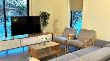 A modern living room with a large flat-screen TV, a grey sofa, two armchairs, a wooden coffee table, and a potted plant.