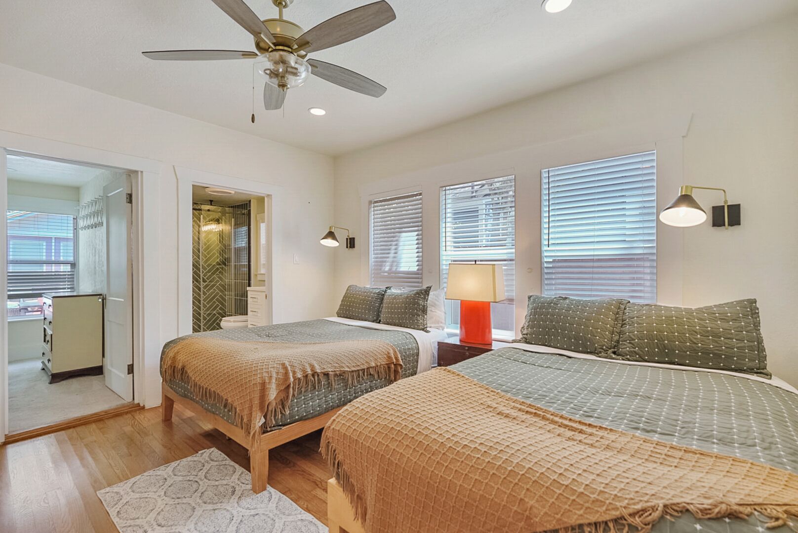 A bedroom with two beds, a ceiling fan, a nightstand with a lamp, and a window with blinds.