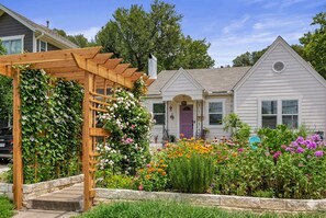 Una casa residencial con una pérgola de madera cubierta de plantas trepadoras y una variedad de flores coloridas en el jardín delantero.
