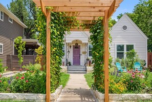 En trädgårdstig som leder fram till ett hus med en lila dörr, omgivet av grönska och blommor.