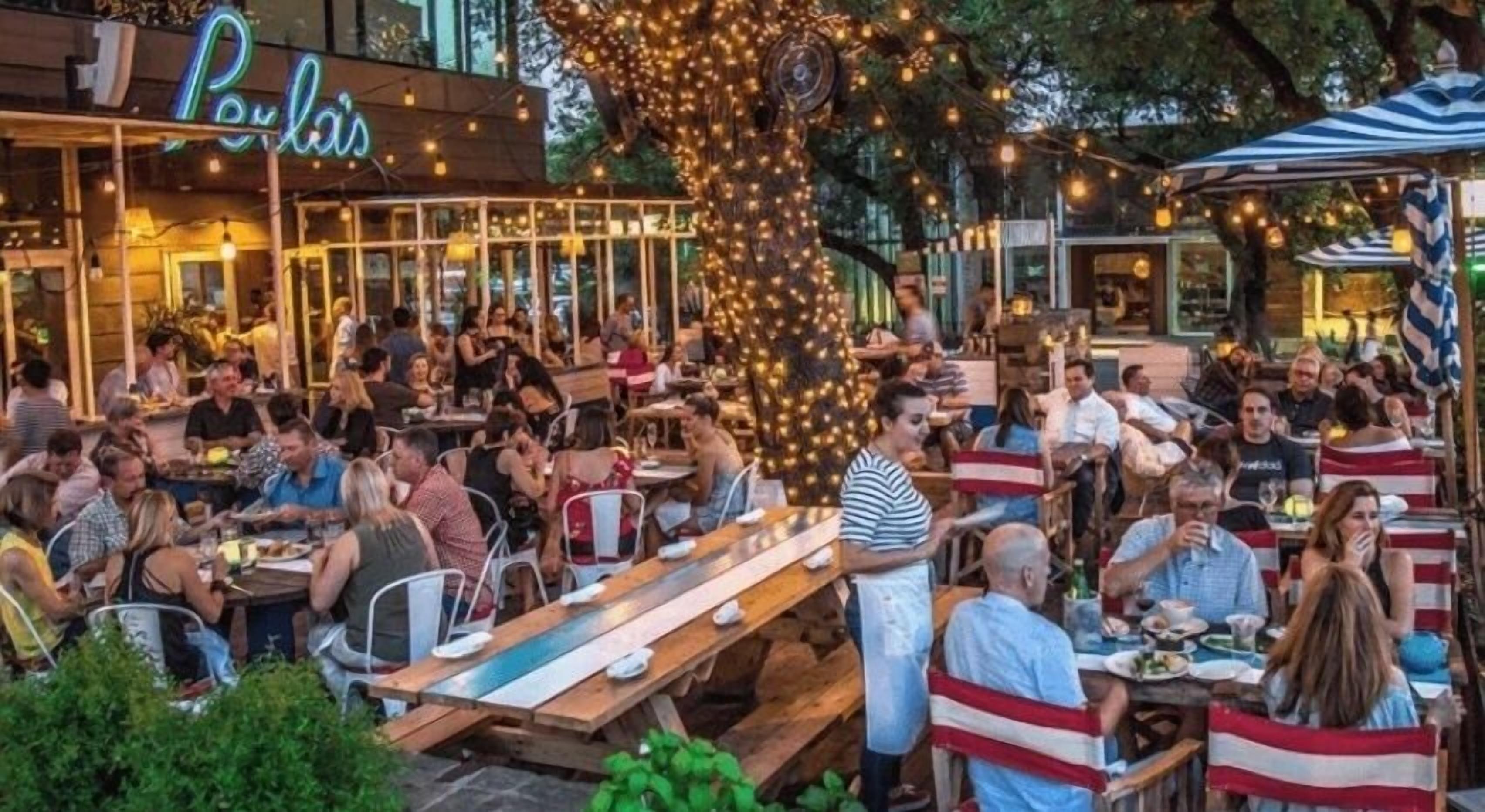 Ein Außenbereich mit Essbereich, Tischen, Stühlen und einem Baum, im Hintergrund das Restaurant 'Poulis'.