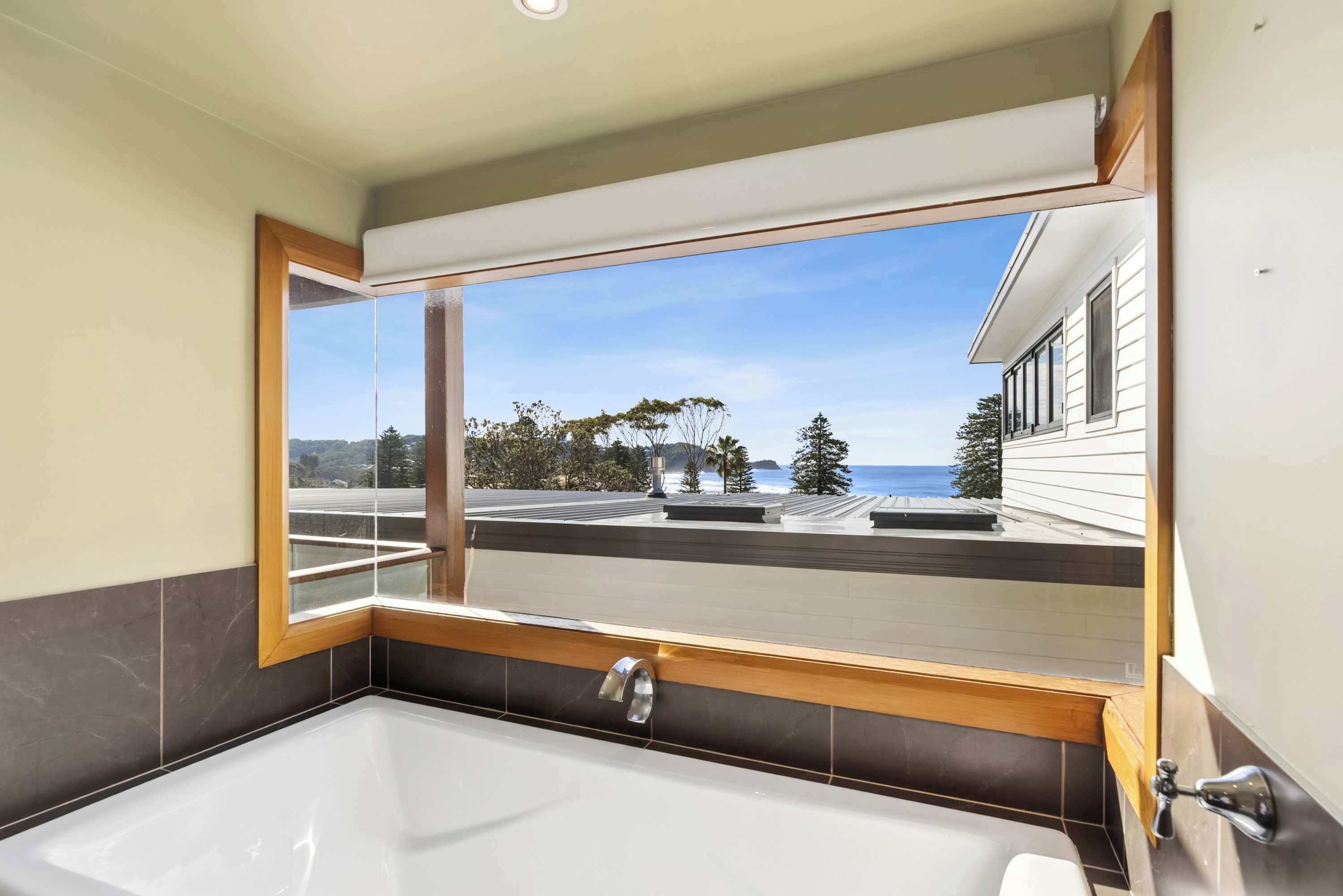 A modern bathroom with a large bathtub, a window with a view of the sea, and a wooden-framed window.