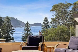 A balcony with wicker chairs, a view of the ocean, and trees. - Avoca Village. 150m to Beach. Sleeps 10 (Avoca Beach)