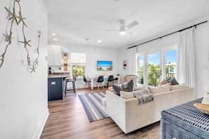 A modern living room with a sofa, a dining area with chairs, a kitchen area, and a balcony with curtains. - Hidden Lake Condos 131, walk to Gulf Place! (Santa Rosa Beach)
