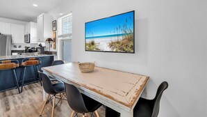 Eine moderne Küche mit weißen Schränken, ein Essbereich mit Holztisch und -stühlen sowie ein Strandbild an der Wand.