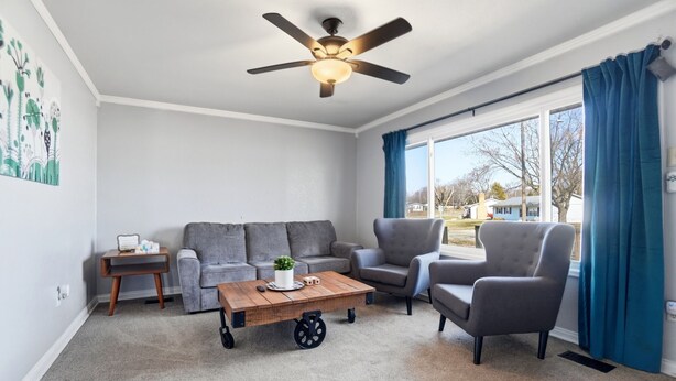 Una sala de estar con ventilador de techo, un sofá gris, una mesa de centro, un sillón gris y vistas al exterior a través de amplios ventanales.