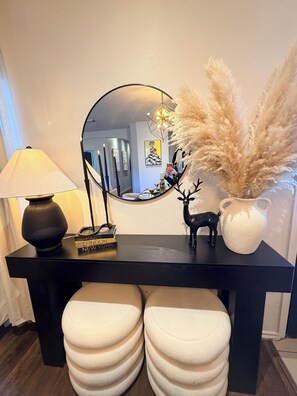 Une console noire surmontée d’un miroir rond, d’une lampe, d’un vase contenant des plumes d’herbe des pampas, d’une petite figurine de cerf et d’un livre intitulé « Paris, Londres, New York ».
