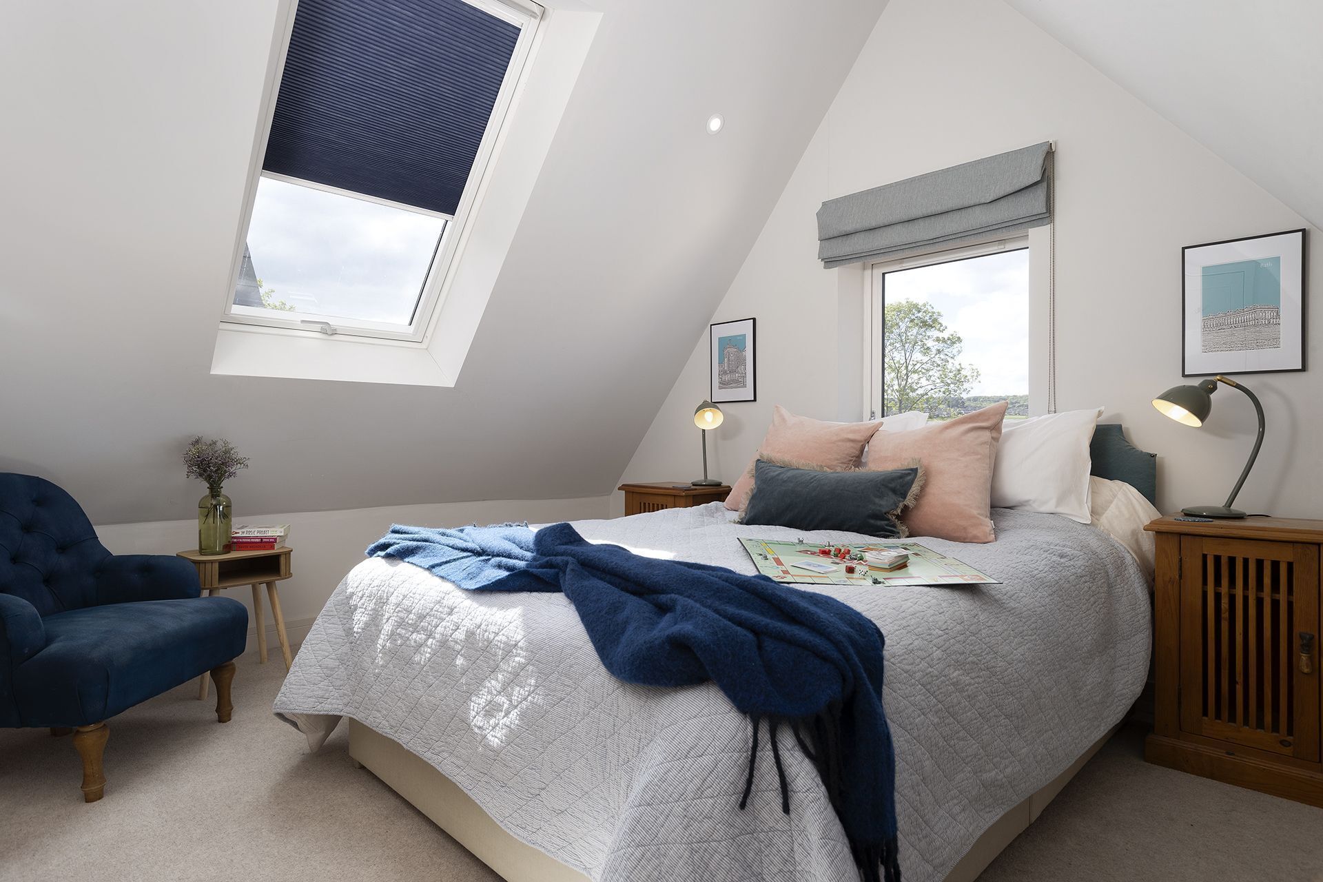 A bedroom with a sloped ceiling, a bed with multiple pillows, a bedside table with a lamp, a chair, and a window with blinds.