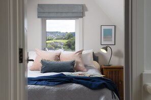 Ein Schlafzimmer mit einem Bett, Kissen, einem Nachttisch mit Lampe, einem Fenster mit Aussicht und einem gerahmten Bild an der Wand.