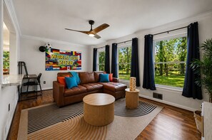 Ein modernes Wohnzimmer mit einer braunen Ledersofa, einem runden Holztisch und einem Deckenventilator. Ein großes Fenster mit dunklen Vorhängen bietet Blick auf Grünflächen außerhalb.