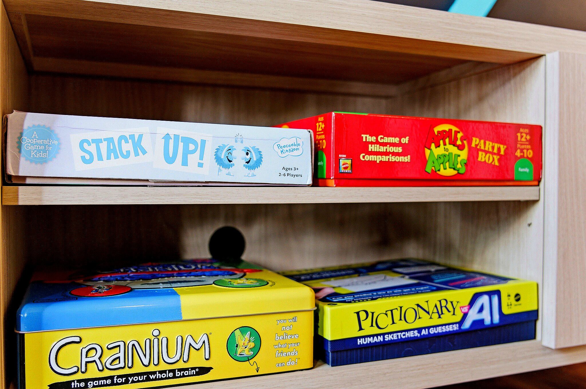 A shelf with board games including 'Stack Up!', 'The Game of Hilarious Comparisons', 'Cranium', and 'Pictionary AI'.
