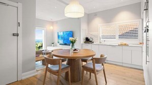 Een moderne keuken met witte kasten, een ronde houten eettafel met stoelen, een hanglamp en een groot raam met jaloezieën.