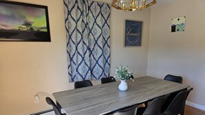 Un coin repas avec une table rectangulaire, des chaises noires, un vase de fleurs, des rideaux à motifs, une citation encadrée et un tableau.