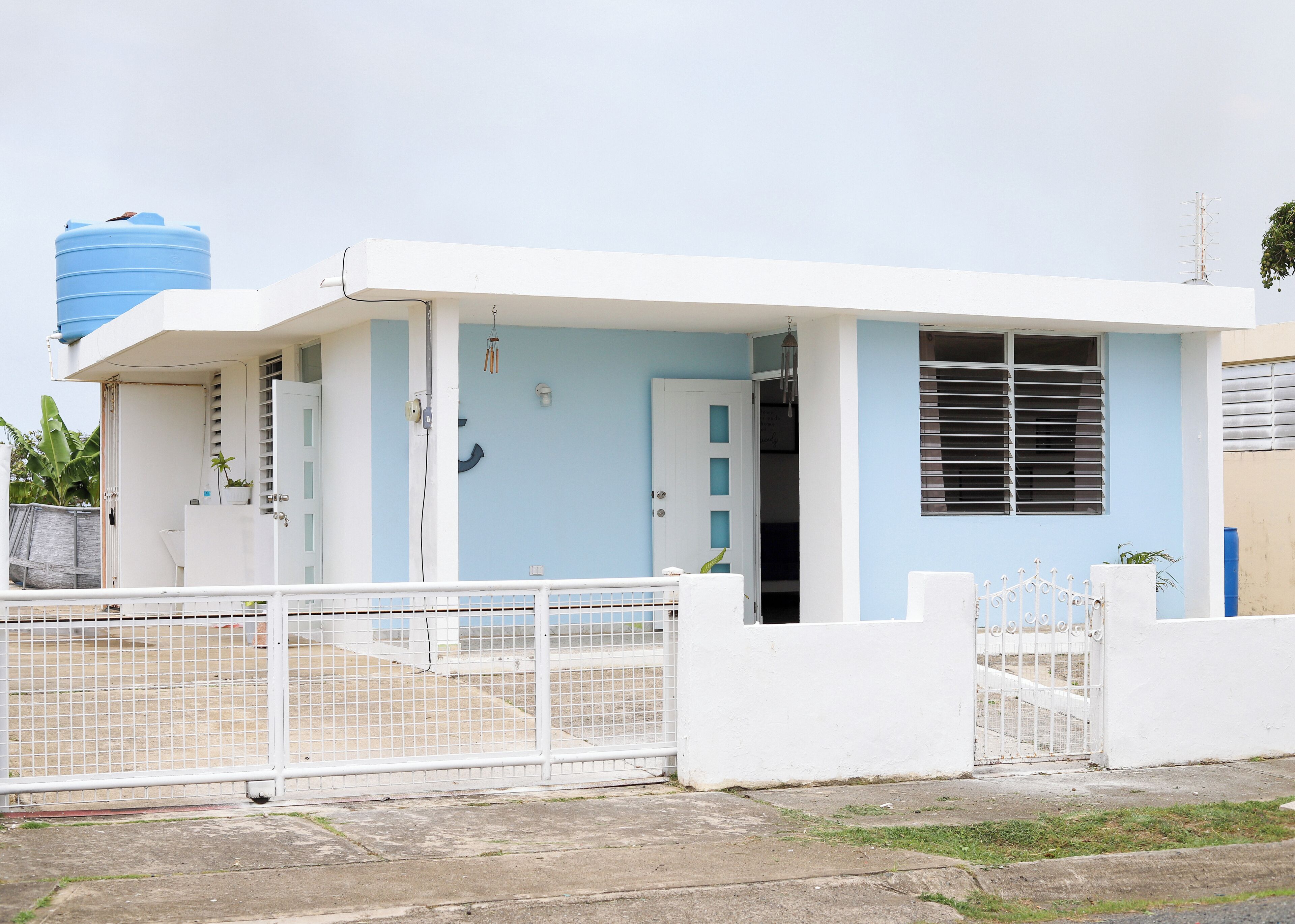 Un bâtiment d’un seul étage, à l’extérieur blanc et bleu clair, avec une porte en métal et un réservoir d’eau bleu en arrière-plan.