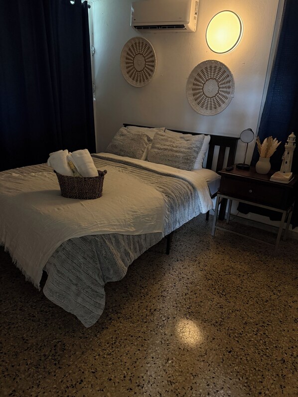 A neatly made bed with a basket of towels, a bedside table with a lamp and a vase, and a wall-mounted air conditioner.