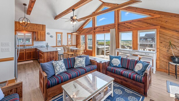 Amplia sala de estar con vigas de madera, cocina y grandes ventanales con vistas al exterior.