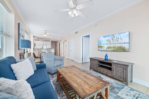 A living room with a blue sofa, a wooden coffee table, a television, and a painting of a beach scene. - Crystal Shores 1307 | Pools + Hot Tub (Gulf Shores)
