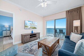A living room with a sofa, coffee table, and a TV stand. There is a balcony with a view of the sky. - Crystal Shores 1307 | Pools + Hot Tub (Gulf Shores)