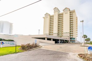 A hotel entrance with a parking area, a sign for Crystal Shores, and a building with multiple balconies. - Crystal Shores 1307 | Pools + Hot Tub (Gulf Shores)