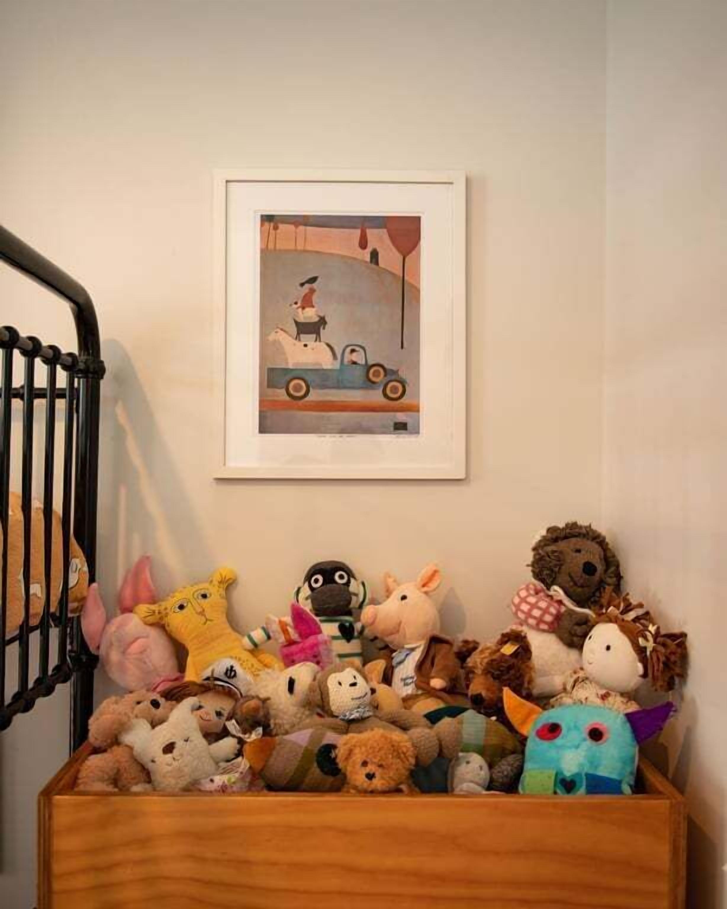 Un berceau en bois avec un bébé à l’intérieur, un mur orné d’un tableau encadré et une pile de peluches.