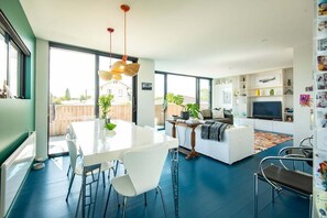 A modern living room with a dining area, large windows, and a TV. - Urban Art House - Modern City Retreat with Spa Pool (Nelson)